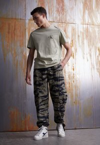 T-shirt en coton vert clair avec un petit logo ; pantalon cargo à motifs en vert et noir ; baskets montantes blanches. Fond texturé.