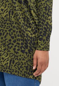 Långärmad topp i olivgrön tyg med ett svart leopardmönster, med rundad nederkant och enkla sömnadsdetaljer.