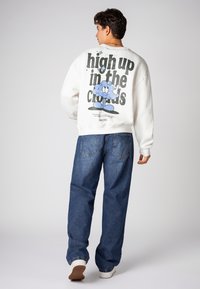 Weißer Sweatshirt mit großem Grafiktext "hoch in den Wolken" und einem Cartoon-Wolken-Design. Kombiniert mit blauen weit geschnittenen Jeans und weißen Sneakers.