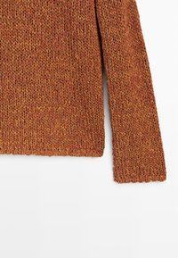 Manche et section du torse d'un pull en grosse maille orange rouille avec un motif tissé texturé sur fond blanc.