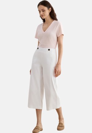 Mujer con top de manga corta en rosa claro con escote en pico, pantalones blancos de pierna ancha recortados con botones, y mocasines beige sin cordones, de pie sobre fondo blanco.