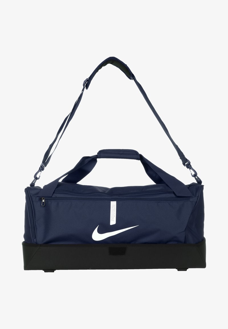 Navy blue duffel bag with a white Nike logo, featuring a zip closure, padded handles, and an adjustable shoulder strap. Black base for durability.
