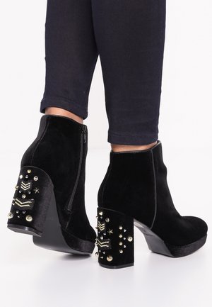 Botas de tobillo de terciopelo negro con un tacón grueso que presenta remaches decorativos y patrones. Cierre de cremallera lateral y un acabado texturizado.