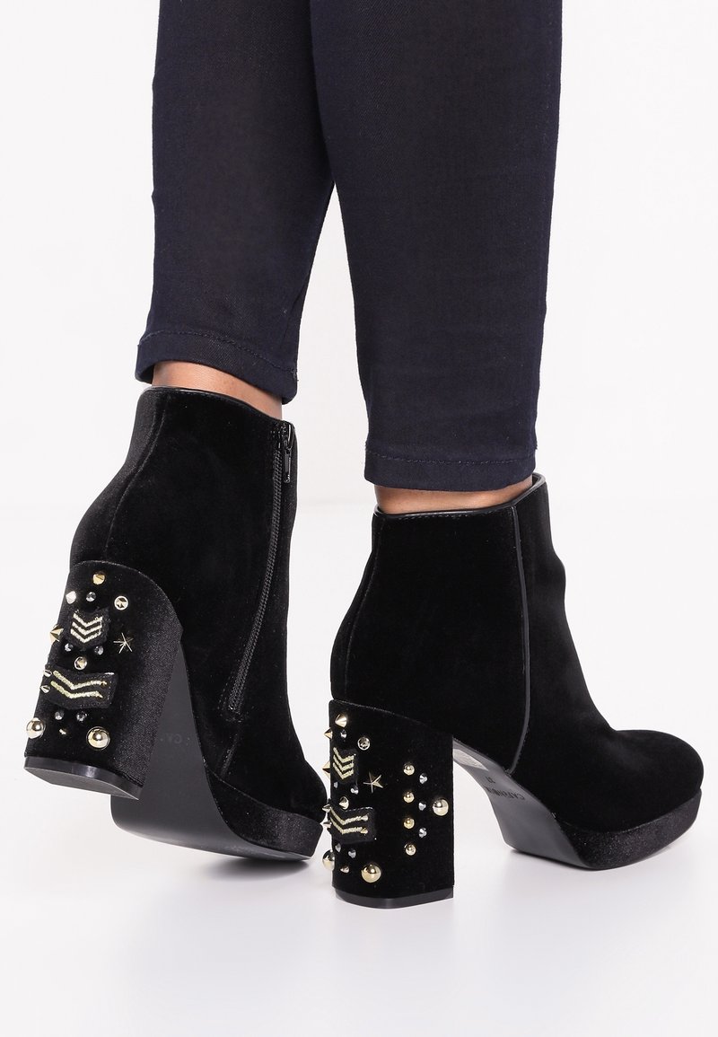 Botas de tobillo de terciopelo negro con un tacón grueso que presenta remaches decorativos y patrones. Cierre de cremallera lateral y un acabado texturizado.