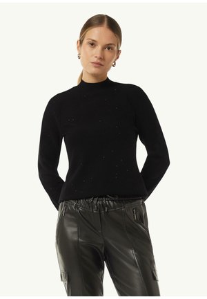 Schwarzer Strickpullover mit hohem Kragen, der dezente Verzierungen aufweist. Getragen mit schwarzen Kunstlederhosen, die Reißverschlussakzente haben.