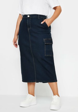 Falda midi de mezclilla en azul oscuro con corte recto, que cuenta con bolsillos laterales y costuras en contraste. Combinada con zapatillas blancas.