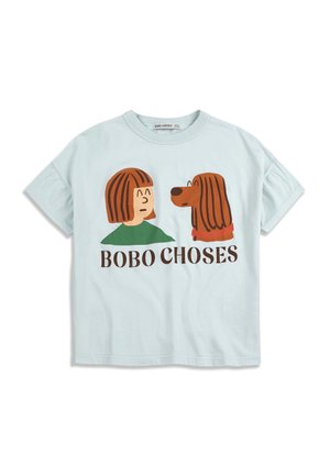 Junges Mädchen trägt ein hellblaues T-Shirt mit Cartoon-Hund und Mädchen-Motiv sowie helle Jeans mit Bauer- und Kuh-Aufnähern, steht mit einer Hand in der Tasche.