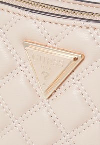 Lichtbeige, gewatteerde handtas met een gouden driehoekig logo met "GUESS" en een vraagteken. Gehaakte oppervlakte met zichtbare stikselaccenten.