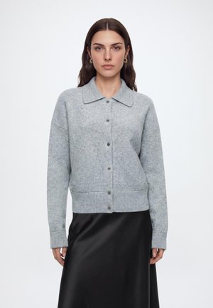 Femme aux longs cheveux foncés porte un cardigan gris boutonné avec col et une jupe en satin noire, debout devant un fond blanc uni.