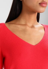 Sweter w kształcie litery V w intensywnym czerwonym kolorze, o żebrowanej fakturze, dopasowanym kroju i długich rękawach, eksponujący gładką linię dekoltu i ramion.