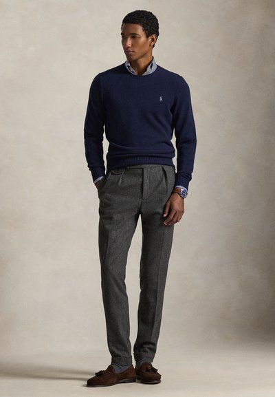 Man draagt een marineblauwe trui over een gestreept overhemd, grijze geplooide broek, bruine loafers met kwastjes en een horloge, staat met handen in de zakken.