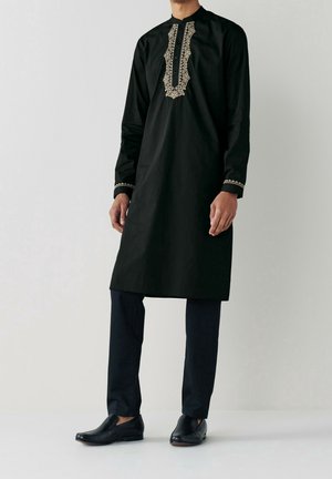 Homme portant une kurta traditionnelle noire avec des détails brodés beige sur le devant et aux poignets, associée à un pantalon noir et des chaussures noires.