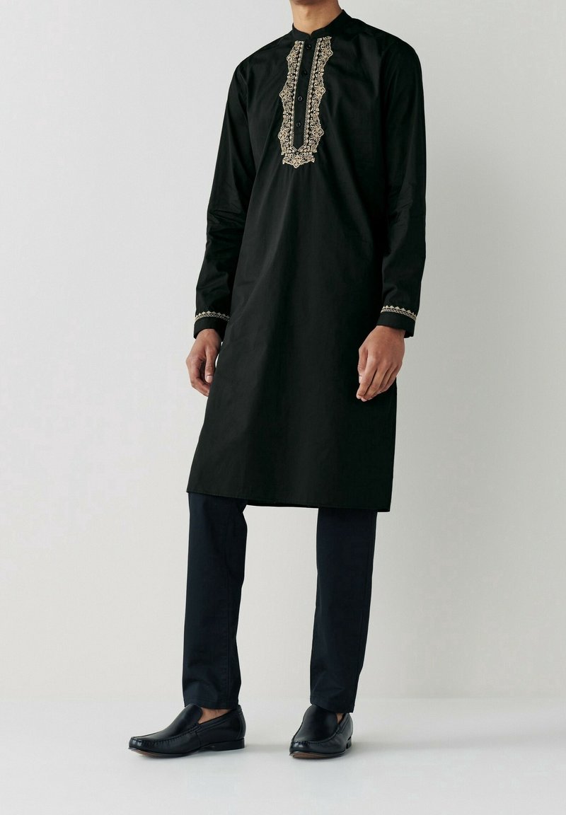 Homme portant une kurta traditionnelle noire avec des détails brodés beige sur le devant et aux poignets, associée à un pantalon noir et des chaussures noires.