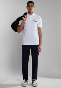 Witte poloshirt met korte mouwen, kraag en geborduurd logo, gecombineerd met donkerblauwe broeken en sneakers, terwijl hij een zwarte tas vasthoudt.