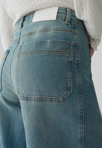 Helles blaue Jeans mit entspannter Passform, ausgestattet mit Hintertaschen und heller Stickerei. An der Taille ist ein weißes Leder-Label sichtbar.