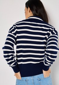 Marineblau und weiß gestreifter Cardigan mit einem gerippten Kragen und Bündchen, der einen lockeren Schnitt und ein strukturiertes Strickgewebe auf einem hellen Hintergrund aufweist.