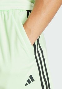 Pantaloni sportivi verde chiaro in tessuto testurizzato, con strisce nere sui lati e un logo ricamato in basso.