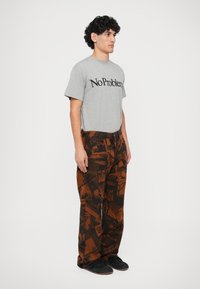 Graues Kurzarm-T-Shirt mit dem Aufdruck "No Problem"; braune Cargo-Hosen mit grafischem Muster; schwarze Schuhe; stehende männliche Figur.