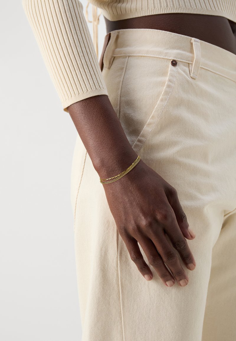 Top a maniche lunghe a coste color crema abbinato a pantaloni beige a vita alta. Al polso è presente un braccialetto multilayer in oro.