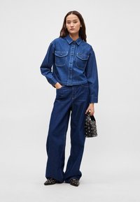 Sötétkék farmerdzseki két elülső zsebbel, gombzárral és hosszú ujjakkal, széles szárú farmernadrággal és fekete szegecselt kézitáskával párosítva.