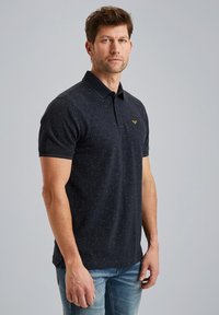Camisa polo negra con un patrón sutil, de manga corta y con un cuello clásico. Detalle del logo dorado en el lado izquierdo del pecho. Combinada con jeans de color azul claro.