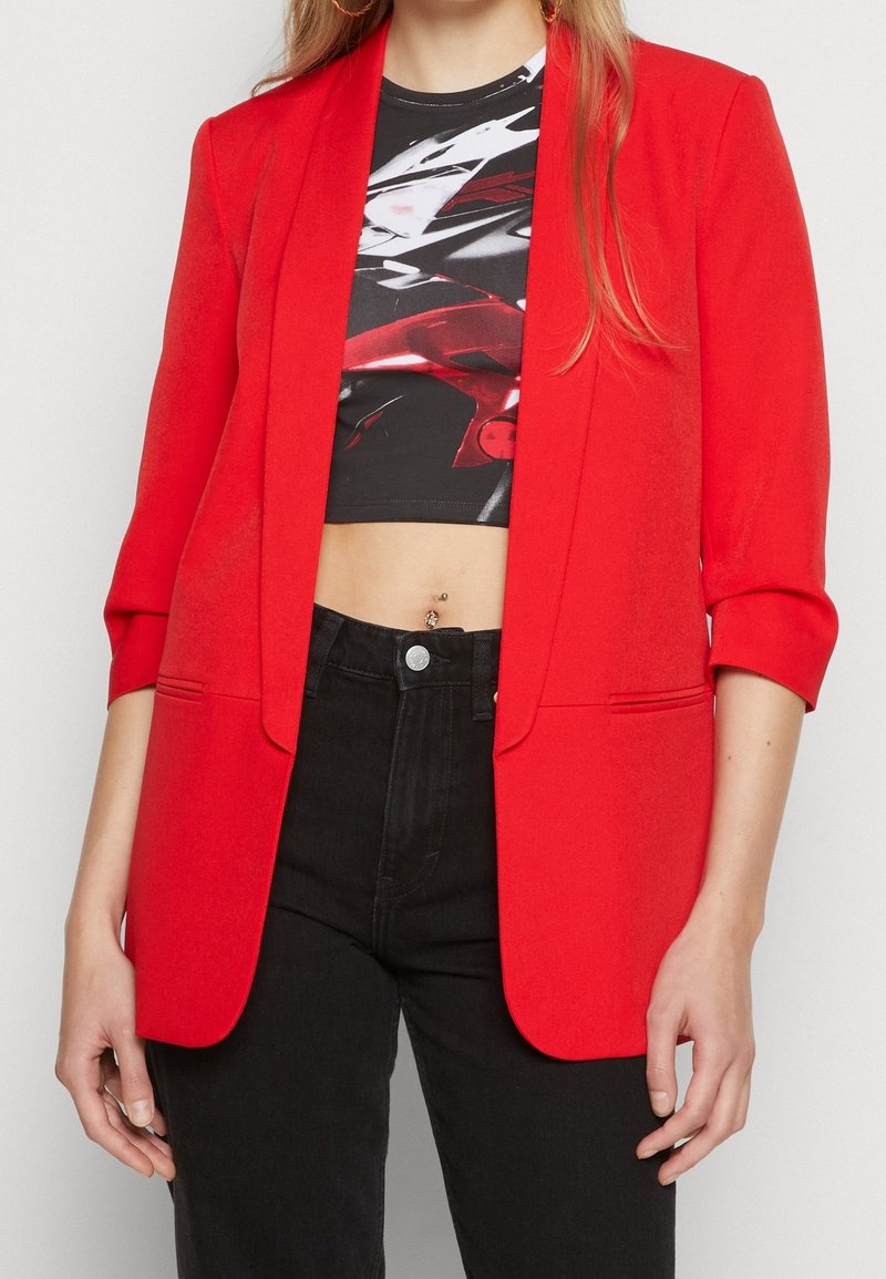 Röd blazer med skräddarsydd passform och notched lapels, med trekvartsärmar. Kombinerad med en svart kort topp och högmidjade svarta jeans.