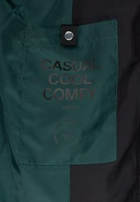Grünes und schwarzes Stoffetikett mit fettem Text, der "CASUAL COOL COMFY" und "S.OLIVER" aussagt, sowie einer Grafik für Pflegehinweise darunter.