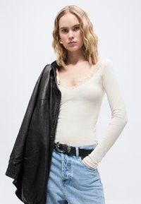 Noor naine laineliste blondide juustega kannab valget pitsivõõrast külgedega topi, helerohelisi teksapükse musta vööga ning hoiab musta nahkjopet õlal.