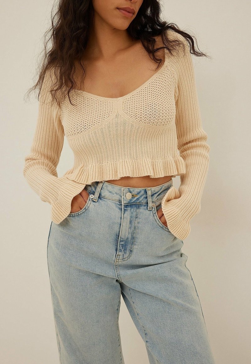 Béžový pletený crop top se širokým výstřihem, žebrovanou strukturou, rozšířenými rukávy a volánovým lemem, kombinovaný se světlými modrými džíny s vysokým pasem.
