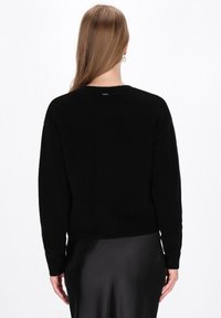 Donna con lunghi capelli castano chiaro, vestita con un maglione nero a maniche lunghe e una gonna nera di raso, di spalle contro uno sfondo bianco.