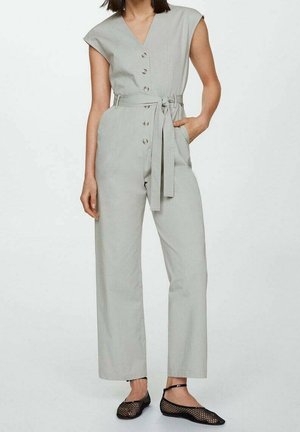 Woman wearing sleeveless light gray buttoned jumpsuit with belt, her right hand in pocket, paired with black mesh flats.