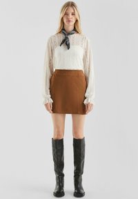 Kobieta ubrana w białą koronkową bluzkę z długim rękawem, brązową zamszową mini spódnicę, czarny szalik bandana oraz czarne skórzane kozaki do kolan, stoi.