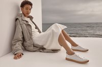 Femme en veste beige, robe en maille blanche et baskets blanches assise par terre près de l'océan sous un ciel couvert.