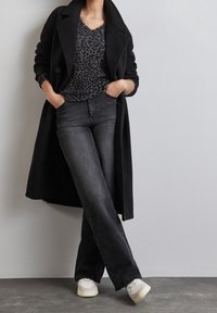 Svart ullkappa över en grå leopardmönstrad tröja, tillsammans med högmidjade svarta jeans och vita sneakers, stående mot en vägg.