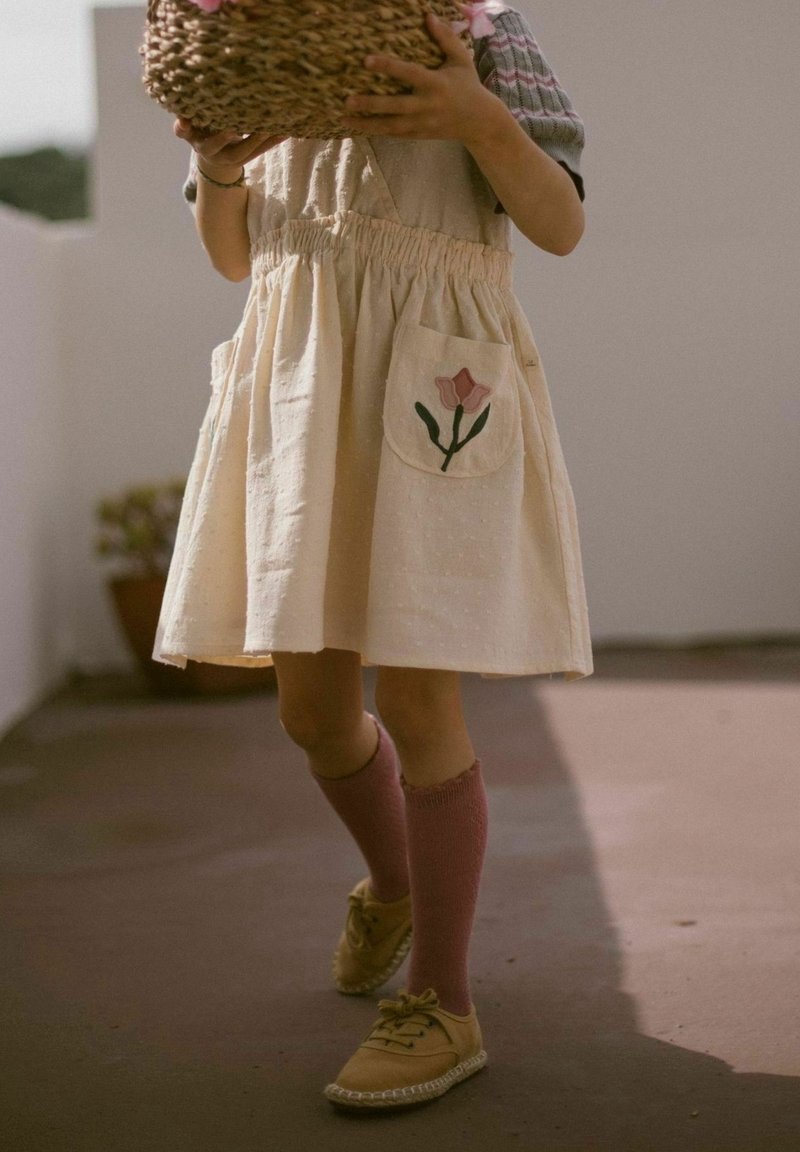 Enfant portant une robe crème avec une poche fleurie brodée, des chaussettes roses montantes et des chaussures jaunes, tenant un panier tressé.