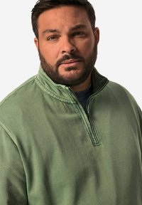 Grön bomullströja med dragkedja i kragen och texturerad yta. Har ett subtilt mönster och avslappnad passform, lämplig för avslappnade tillfällen.