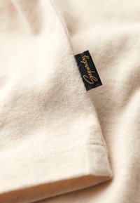 Lichtbeige katoenen stof met een gestructureerde afwerking, met een zwart label met gouden letters dat aan de rand is bevestigd.