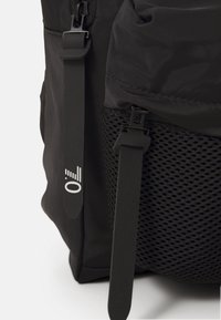 Zaino nero con pannello in rete e spallacci neri regolabili. Presenta un marchio sottile in bianco. Tessuto testurizzato con accenti lisci.