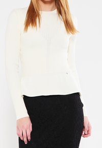 Cremfarbener, gerippter Langarm-Top mit taillierter Passform, V-förmigem Design und Volant-Saum, kombiniert mit einem schwarzen, strukturierten Rock.