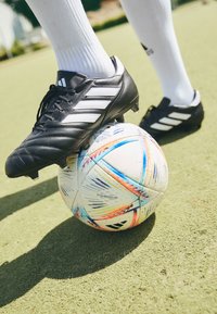 Botín de fútbol negro con tres franjas blancas descansando sobre un balón de fútbol multicolor con patrones visibles, sobre una superficie parecida a la hierba.