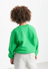 Maglione verde con collo rotondo e maniche raglan, realizzato in tessuto morbido. Pantaloni grigi con tasca e un accento giallo.