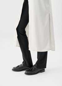 Μαύρα loafers με φουντίτσες σε συνδυασμό με στενά μαύρα παντελόνια και ένα μακρύ λευκό παλτό με πλαϊνές σκισιές, δημιουργώντας ένα πολυεπίπεδο στυλ.