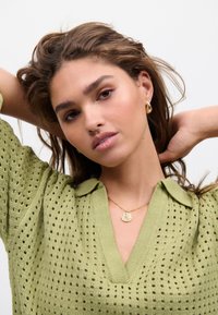 Maglione verde lavorato a maglia con scollo a V e tessuto a rete, abbinato a una collana con pendente d'oro e piccoli orecchini a cerchio.