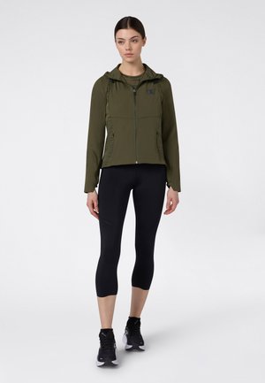 Femme debout portant une veste zippée à capuche olive, un legging court noir et des chaussures de sport noires sur un fond uni.