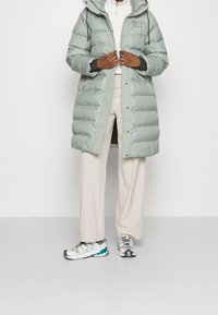 Uppblåsbar jacka i ljusgrön med huva, med dragkedjestängning och ribbade ärmslut. Bärs med vita byxor och flerfärgade sportskor.