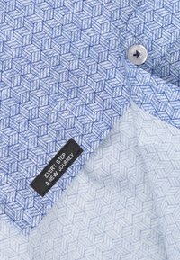 Blauw patroonstof met geometrische vormen en een subtiele textuur. Inclusief een witte knop en een zwart label met witte tekst.