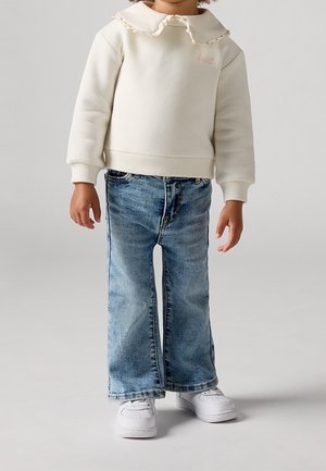 Kind draagt een gebroken witte trui met gerimpelde kraag, lichtblauwe wijde spijkerbroek en witte sneakers, staand tegen een effen achtergrond.