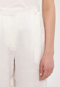 Bílé kalhoty s jemným květinovým vzorem, lehký materiál, vysoký pas a boční kapsy; obsahují stuhu jako ozdobu na pase.