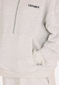 Hellgrauer Sweatshirt mit Reißverschlusskragen, Vorderseite mit Tasche, gerippten Bündchen und einem aufgestickten Logo "LES'ARCS" in Schwarz. Weicher Stoff.