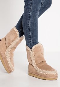 Beige semskete ankle støvler med dekorativ sømkant, myk tekstur og flat såle, kombinert med mørkeblå skinny jeans.